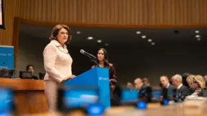Moteris šviesiu kostiumėliu kalba prie mėlynos tribūnos oficialiame susitikime su auditorija fone.