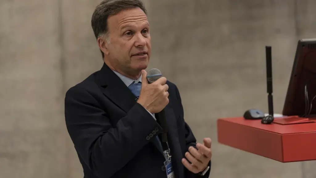 Vidutinio amžiaus vyras su kostiumu ir mikrofonu kalba auditorijai konferencijos metu.