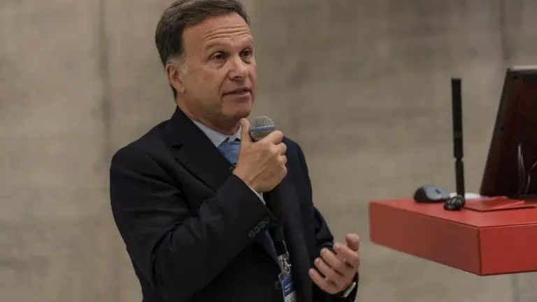 Vidutinio amžiaus vyras su kostiumu ir mikrofonu kalba auditorijai konferencijos metu.
