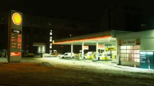 Degalinė naktį su šviečiančiu kainų stendu ir snieguotu privažiavimu automobiliams.
